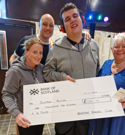 Four people hold a giant donation cheque, indoors, disco lights