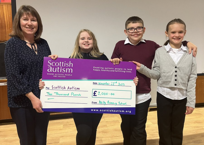Image of an adult and three children holding a giant cheque, in a school, Text on cheque reads 'Scottish Autism, Two thousand pounds, November 22nd 2023, From Kelty Primary School'