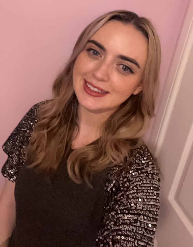 Image of a person wearing a sparkly black top standing infront of a pink wall, indoors