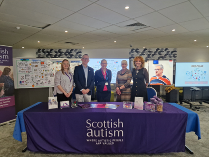 Image is of people standing behind an information table at an event, the table has Scottish Autism branding all around it